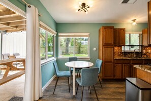 A kitchen with wooden cabinets, a round table with chairs, and a view of the backyard through large windows. - Cozy Home | Pool Table & 2 Arcade Games | Enclosed Backyard (San Antonio)