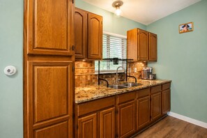A kitchen with wooden cabinets, a granite countertop, a sink, and a window with blinds. - Cozy Home | Pool Table & 2 Arcade Games | Enclosed Backyard (San Antonio)