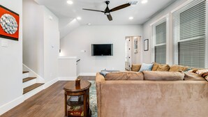 A modern living room with a large beige sofa, a wooden side table, a ceiling fan, and a flat-screen TV mounted on the wall.