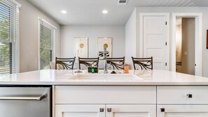 A modern kitchen with white cabinetry, a stainless steel refrigerator, and a white countertop. There are three chairs around a central island with a sink.