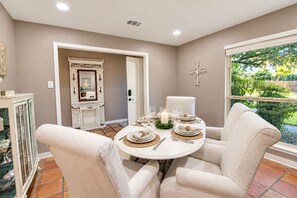 A dining area with a round table set for four, a white chair, and a window with a view of greenery. - Dog-friendly chic home w/private outdoor pool, gas grill, & enclosed backyard (San Antonio)
