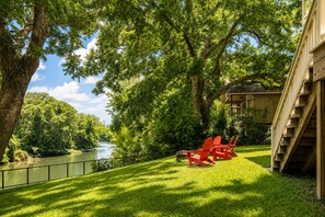 Een pand aan de rivier met een houten terras, rode Adirondack-stoelen en een trap naar een gebouw.