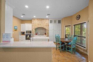 A kitchen with a stone fireplace, white cabinets, a dining table with chairs, and a view through the window.