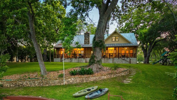 A two-story house with a stone foundation, a large tree in the front yard, and a wooden deck with a bench. - Winter Texan riverfront home on Lake Seguin with dock, kayaks, beautiful views & air hockey (Seguin)