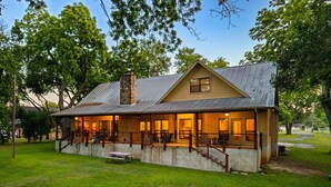 A two-story house with a porch, a stone chimney, and a grassy yard. - Winter Texan riverfront home on Lake Seguin with dock, kayaks, beautiful views & air hockey (Seguin)