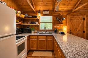 A cabin-style kitchen with wooden cabinets, granite countertops, a white refrigerator, a stove, a sink, and a window with blinds. - Modern Hill Country Multi-unit w/kitchen, WIFI, patio, private pool & hot tubs (Wimberley)