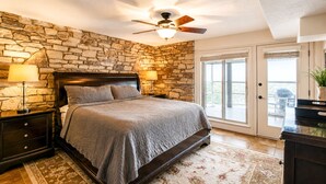 A bedroom with a stone wall, a ceiling fan, a bed with a headboard, bedside lamps, a nightstand, a window with a view, and a door.