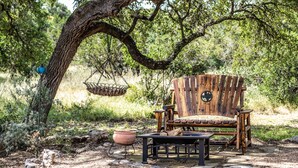 A rustic outdoor seating area with a wooden chair and table, surrounded by natural vegetation and a hanging basket. - The All-Suite Century Oak House - Steps to Remi's Ridge & Hayes Hollow (Spring Branch)