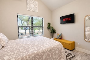 A bedroom with a bed, a TV mounted on the wall, a window with blinds, a plant, and a yellow bench. - Tanglewood Retreat | Updated house in Spring Branch with a large yard (Spring Branch)