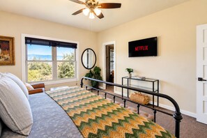 Ein Schlafzimmer mit einem Bett, einem Deckenventilator, einem Fenster mit Blick, einem an der Wand montierten Fernseher und einer Tür.