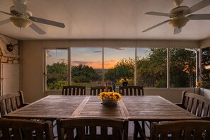 Uma área de jantar com mesa e cadeiras, um ventilador de teto e janelas amplas com vista para árvores e um pôr do sol.