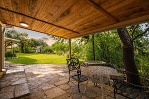 A covered patio with stone flooring, a table, and chairs, overlooking a grassy yard and trees. - Cozy 3BR in Canyon Lake | Grill | Private Backyard and Winter Texan Friendly (Canyon Lake)