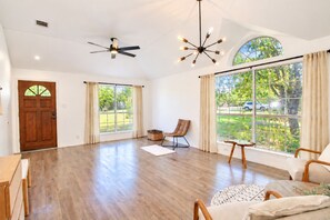A spacious room with hardwood flooring, a ceiling fan, a modern chandelier, and large windows offering a view of the outdoors. - Beautiful dog-friendly home with fireplace, large yard, hammock, W/D, and AC (Buda)