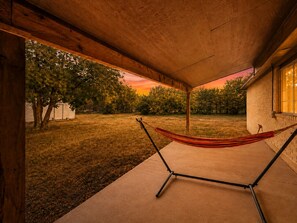 A hammock under a wooden ceiling, a brick wall, and a view of a grassy yard with trees.
