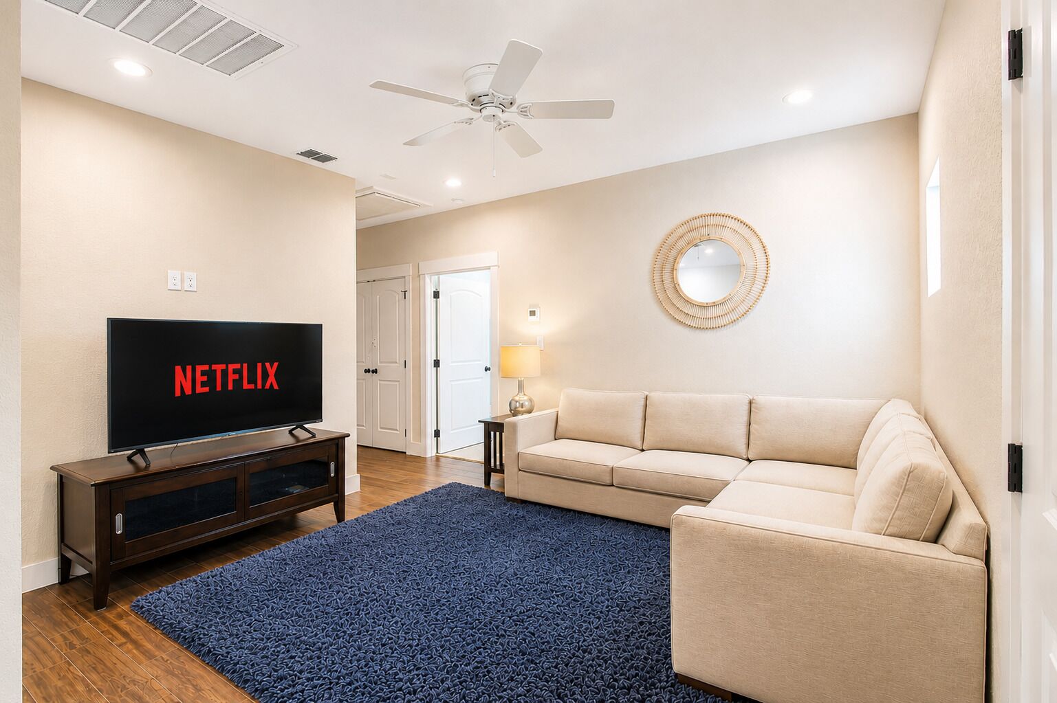 A living room with a television displaying 'NETFLIX,' a ceiling fan, a beige sofa, a blue rug, and a wall-mounted mirror.