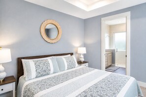Une chambre à coucher avec un lit, deux tables de chevet avec des lampes, un miroir fixé au mur et une salle de bain visible à travers une porte ouverte.