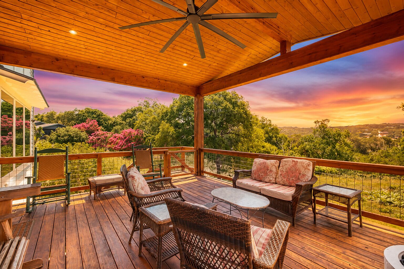 A covered outdoor deck with wicker furniture, a ceiling fan, and a scenic view of trees and hills.
