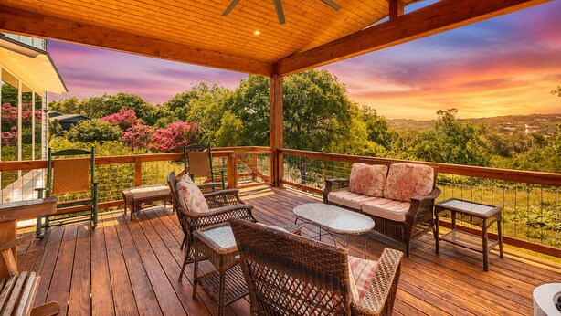 Une terrasse extérieure couverte, aménagée avec des meubles en osier, un ventilateur de plafond et offrant une vue pittoresque sur les arbres et les collines.