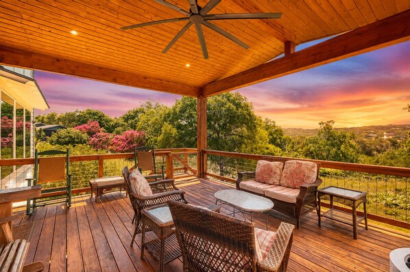 Une terrasse extérieure couverte, aménagée avec des meubles en osier, un ventilateur de plafond et offrant une vue pittoresque sur les arbres et les collines.