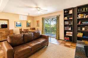 A living room with a brown leather sofa, a wooden coffee table, a TV, a bookshelf, and a sliding glass door to a balcony.