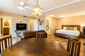 A room with a wooden dining table, a sofa, a bed with a wooden headboard, a TV, and a ceiling fan.