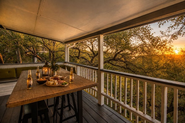 An outdoor wooden deck with a table set for a meal, surrounded by trees and a sunset view.
