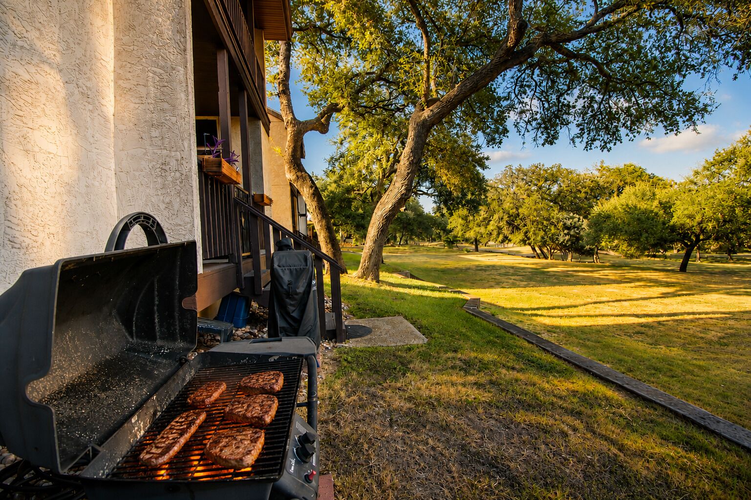 A barbecue grill with food being cooked, a covered porch, and a grassy yard with trees.