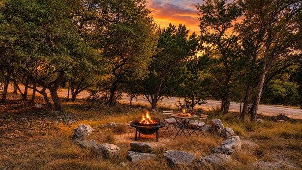 Een kampvuur met een tafel en stoelen in een natuurlijke omgeving, met bomen en een zonsondergang op de achtergrond.