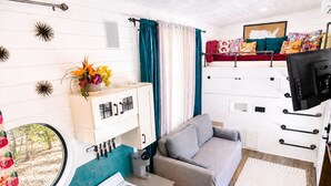 Een ruimtelijk ingerichte leefruimte met kitchenette, een bank, een aan de muur bevestigde televisie en een raam met gordijnen.