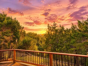 Una terraza de madera con barandilla, rodeada de frondosa vegetación y un cielo al atardecer.
