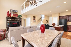 A spacious living room with a high ceiling, a dining area with a marble table, a kitchen with dark cabinetry, and a staircase leading to an upper level.