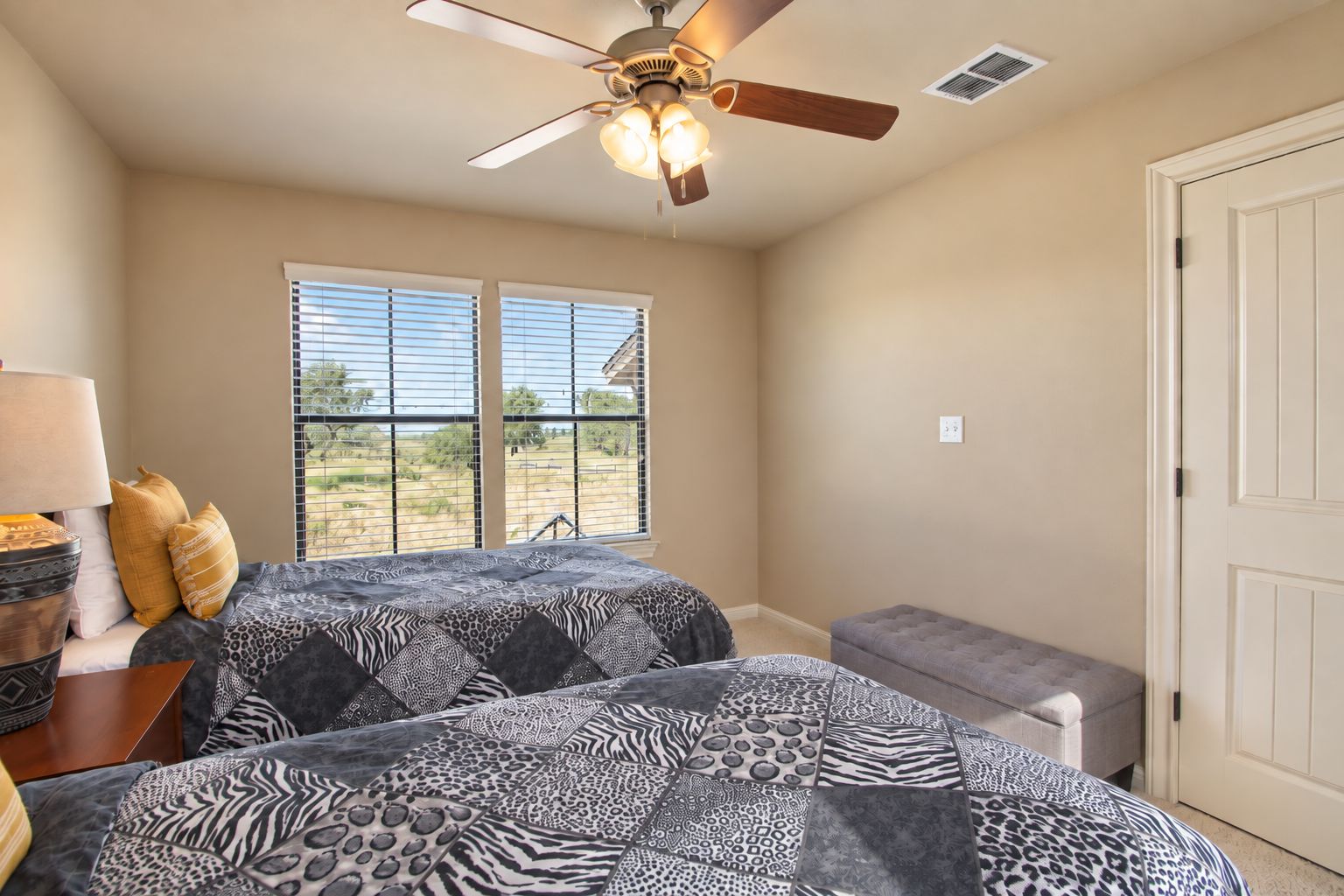 A bedroom with two beds, a ceiling fan, a window with blinds, and a door.