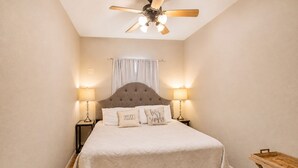 A bedroom with a tufted headboard, two bedside lamps, a bed with a white bedspread, and a ceiling fan. - Gruene Envy – a first floor Poolside Condo with Guadalupe River Access (New Braunfels)