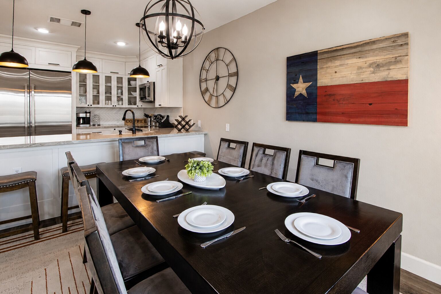Une salle à manger avec une longue table dressée pour six personnes, une cuisine en arrière-plan et un drapeau du Texas accroché au mur.