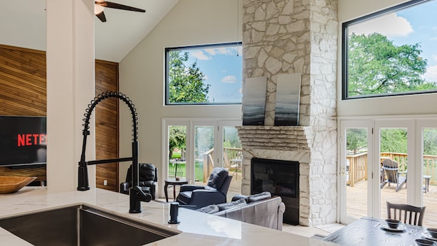 A modern kitchen with a stone fireplace, a dining area with a table and chairs, and a large window overlooking a deck. - Idyllic waterfront getaway with private hot tub & deck views - near golf (New Braunfels)