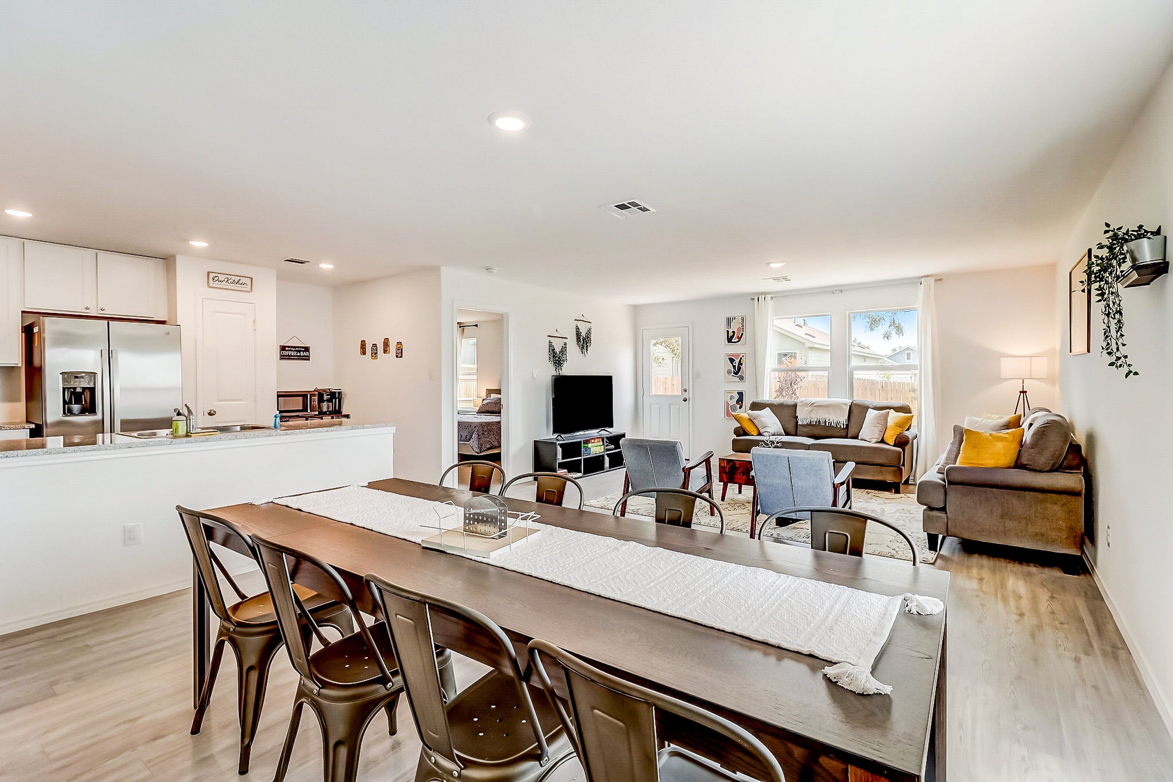 Une cuisine moderne avec des meubles blancs, un coin repas avec une table et des chaises en bois, et un salon avec un canapé et un téléviseur.