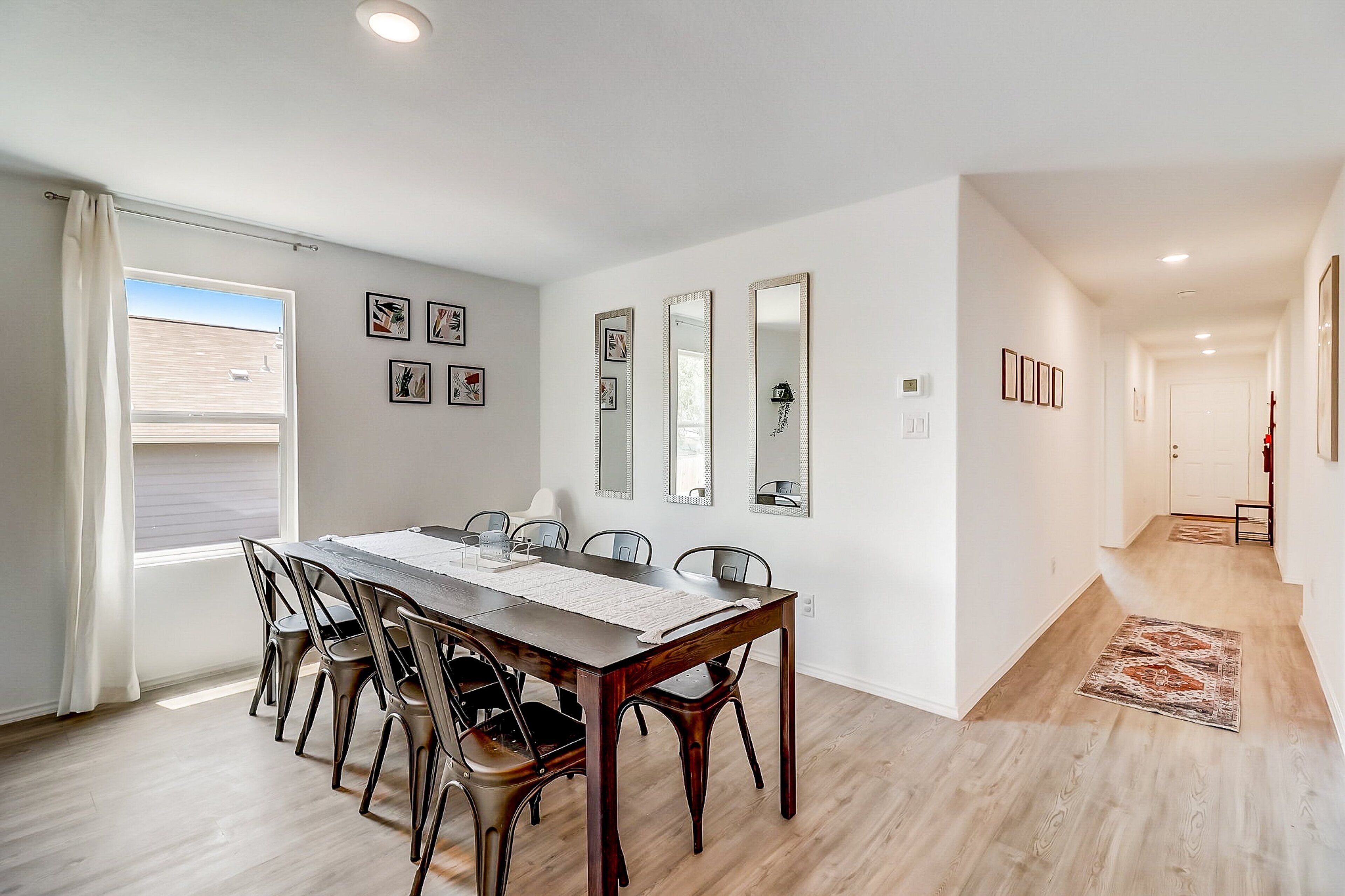 Un área de comedor con una mesa y sillas de madera, una ventana con cortinas, y un pasillo con alfombra.