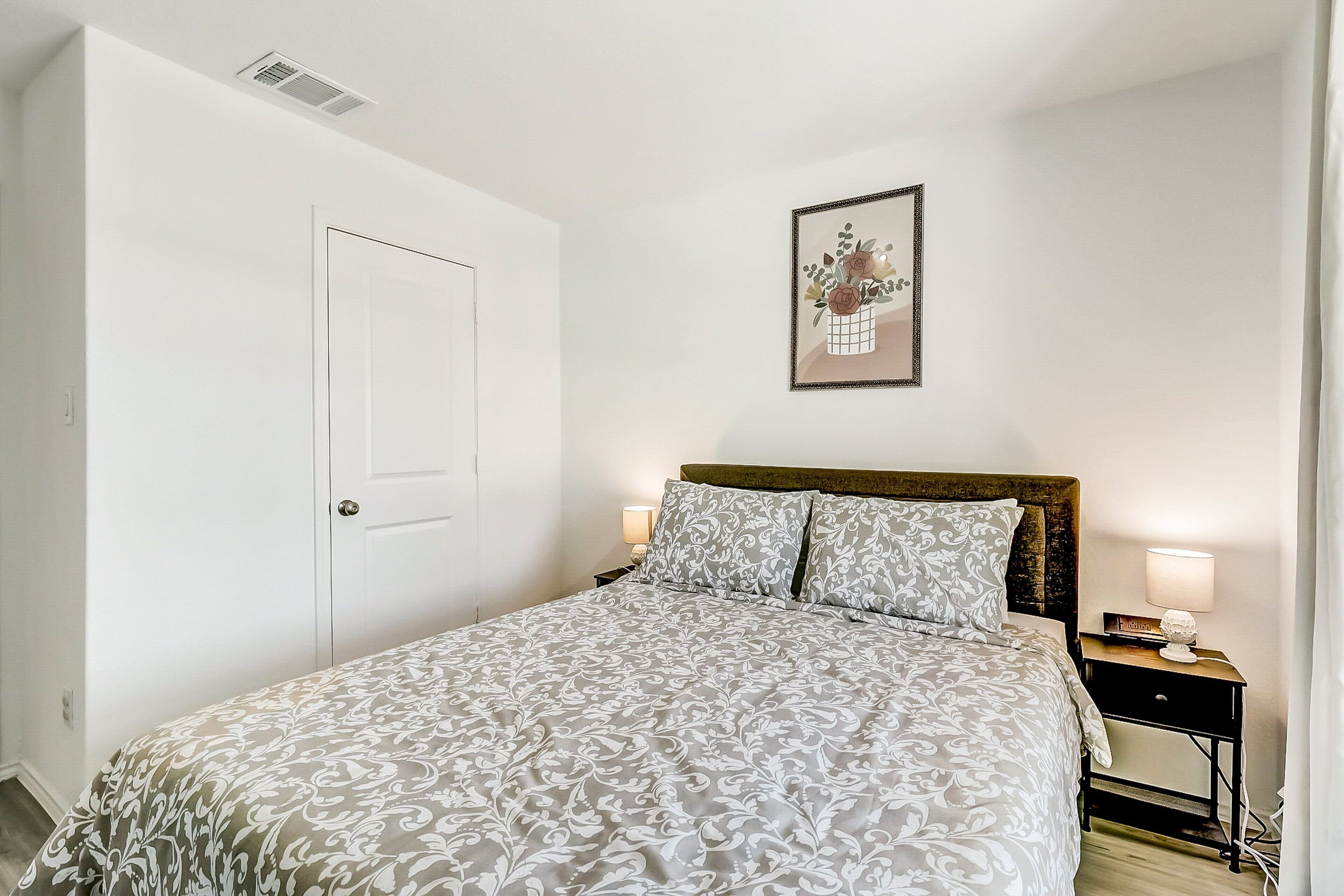 Un dormitorio con cama, mesita de noche, lámpara, un cuadro en la pared y una puerta.