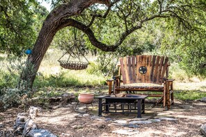A rustic outdoor seating area with a wooden chair and table, surrounded by natural vegetation and a hanging basket. - Walk to Remi's Ridge & Hayes Hollow Century Oak Casita - Near Canyon Lake (Spring Branch)