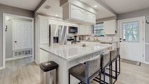 A modern kitchen with white cabinetry, a central island with bar stools, stainless steel appliances, and recessed ceiling lighting. - Stunning Rivrefront 3BR with Hot tub, water view! (Seguin)