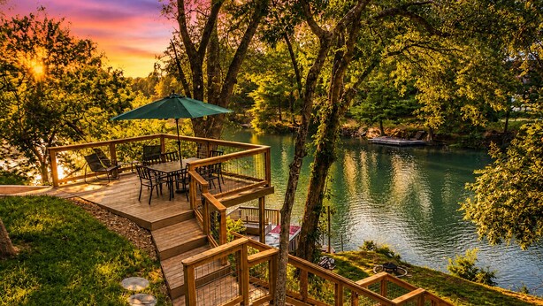 A wooden deck with outdoor furniture, a green umbrella, and a view of a lake surrounded by trees. - Stunning Rivrefront 3BR with Hot tub, water view! (Seguin)