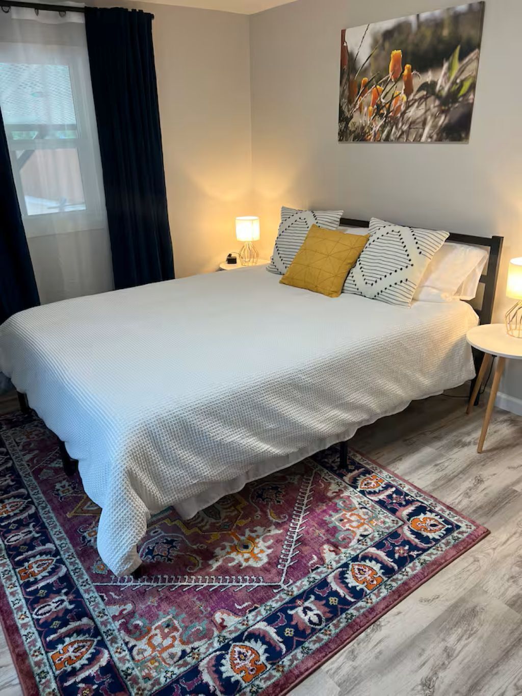 Een net opgemaakt bed met een beddengoed met patroon, een decoratief vloerkleed en een kunstwerk aan de muur boven het bed.