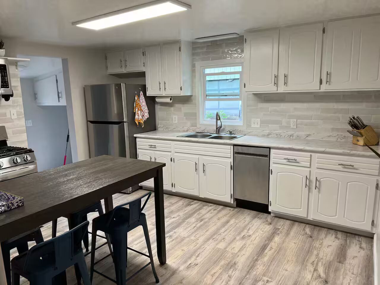 A modern kitchen with white cabinets, a stainless steel refrigerator, a built-in dishwasher, and a wooden dining table with two chairs.