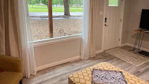 A room with a large window, a door, a small table, and a TV stand. - Remodeled home across from river, train and park! (Cañon City)