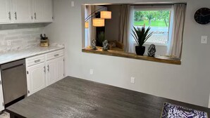A kitchen with white cabinets, a marble countertop, a built-in window with a view, and a dining area with a wooden table. - Remodeled home across from river, train and park! (Cañon City)