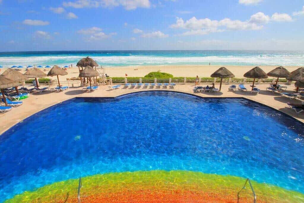 A pool with clear blue water, surrounded by lounge chairs and thatched umbrellas, with a beach and ocean view in the background.