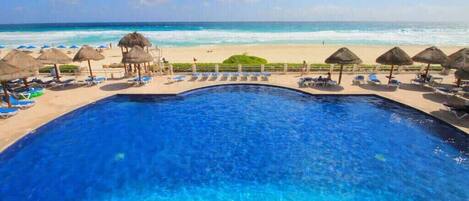 A pool with clear blue water, surrounded by lounge chairs and thatched umbrellas, with a beach and ocean view in the background.