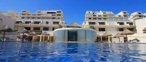 A swimming pool with a clear blue water surface, surrounded by modern buildings with balconies and a central structure with a curved facade.