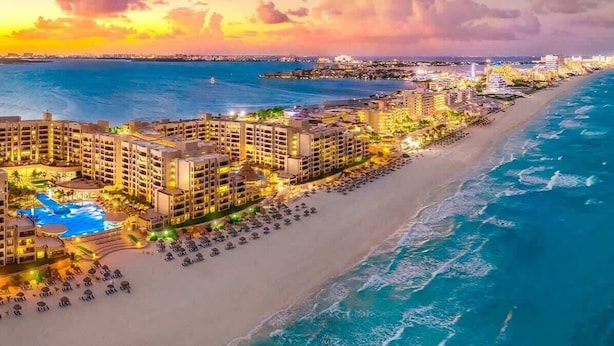Un hotel frente a la playa con varias construcciones, una piscina y una playa con sombrillas.