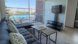 Ein Wohnzimmer mit einer Couch, einem Fernseher und einem Balkon mit Blick auf einen Pool und Gebäude.
